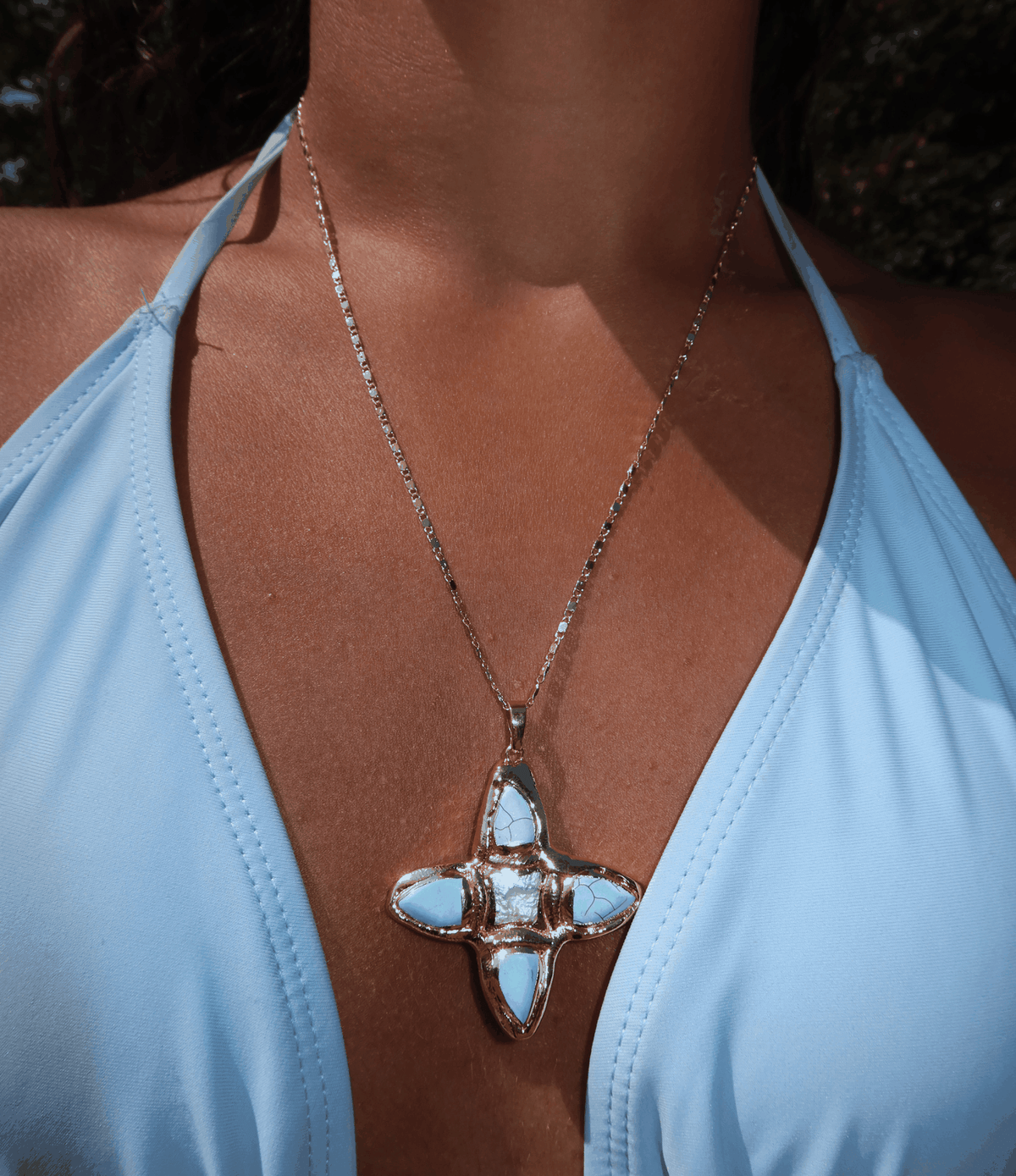 OPHELIA NECKLACE IN BLUE, gold-tone chain with a pale blue stone cross pendant featuring a polished metal setting